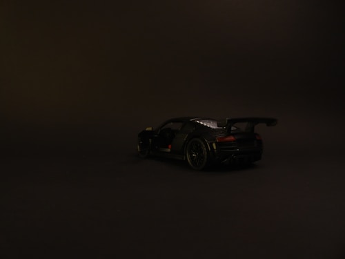 A dark, sleek sports car is positioned against a plain black background, highlighting its aerodynamic design and smooth curves. The lighting is minimal, casting subtle shadows and emphasizing the car's polished surface.
