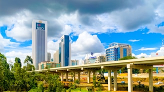 A bustling urban scene featuring modern high-rise buildings, including a prominent skyscraper labeled 'NBO'. These structures are set against a dynamic sky with both bright white and dark gray clouds. In the foreground, there is an elevated highway with several vehicles, and lush green trees add a natural element to the cityscape.