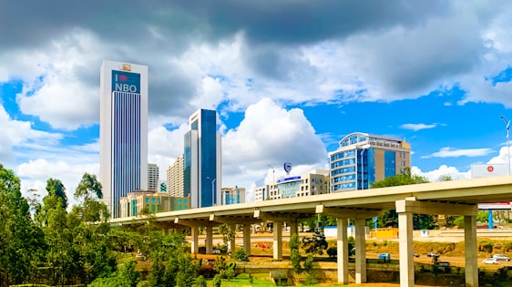 A bustling urban scene featuring modern high-rise buildings, including a prominent skyscraper labeled 'NBO'. These structures are set against a dynamic sky with both bright white and dark gray clouds. In the foreground, there is an elevated highway with several vehicles, and lush green trees add a natural element to the cityscape.