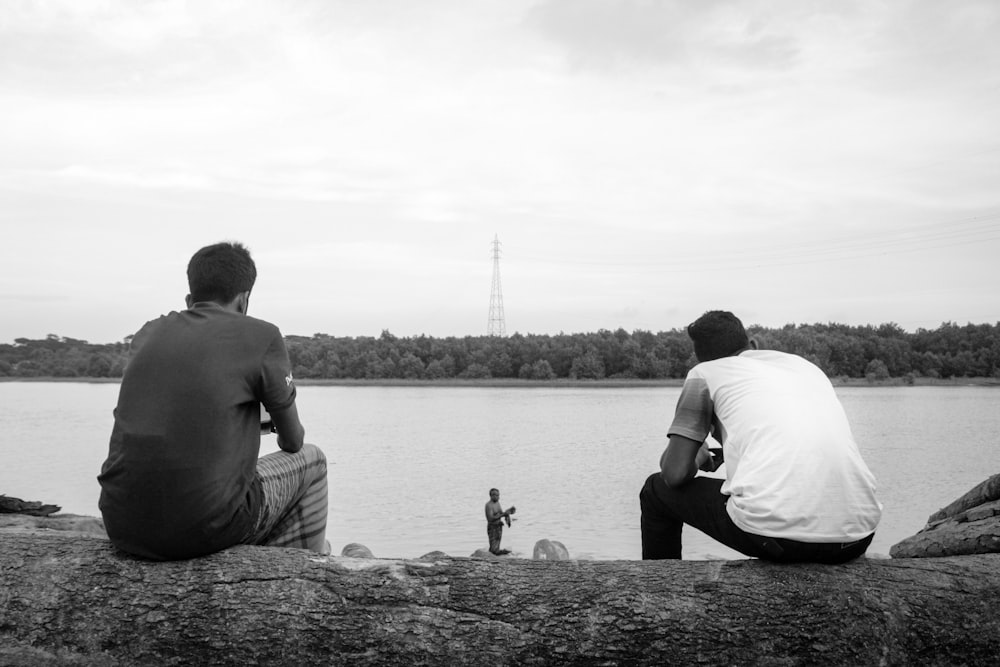 two men sitting on a rock looking at a body of water
