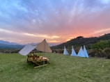 Tents are set up on a grassy field with a scenic backdrop of hills during a vibrant sunset. A wooden picnic table is arranged under one of the tents, while three smaller teepee-style tents stand nearby. The sky is painted with hues of pink, orange, and blue.