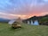 Tents are set up on a grassy field with a scenic backdrop of hills during a vibrant sunset. A wooden picnic table is arranged under one of the tents, while three smaller teepee-style tents stand nearby. The sky is painted with hues of pink, orange, and blue.