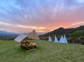 Tents are set up on a grassy field with a scenic backdrop of hills during a vibrant sunset. A wooden picnic table is arranged under one of the tents, while three smaller teepee-style tents stand nearby. The sky is painted with hues of pink, orange, and blue.