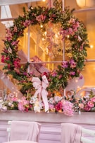A festive holiday wreath hangs on a window, adorned with pink and red flowers and delicate lights. Below the wreath, a floral arrangement with roses and other pink blooms decorates the window ledge. Soft, warm lighting from a chandelier inside creates a cozy and inviting atmosphere.