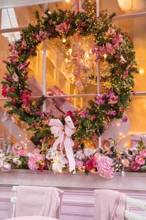 A festive holiday wreath hangs on a window, adorned with pink and red flowers and delicate lights. Below the wreath, a floral arrangement with roses and other pink blooms decorates the window ledge. Soft, warm lighting from a chandelier inside creates a cozy and inviting atmosphere.
