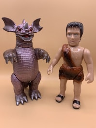 Two toy figures are standing side by side on a plain beige background. The left figure resembles a reptilian monster with large ears, red eyes, and a horn on its nose. Its body is textured like scales and has large, claw-like hands and feet. The right figure is a caveman with a short black hairstyle, a detailed face, and is wearing a faux fur outfit. He has bare feet with black sandals.