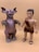 Two toy figures are standing side by side on a plain beige background. The left figure resembles a reptilian monster with large ears, red eyes, and a horn on its nose. Its body is textured like scales and has large, claw-like hands and feet. The right figure is a caveman with a short black hairstyle, a detailed face, and is wearing a faux fur outfit. He has bare feet with black sandals.
