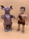 Two toy figures are standing side by side on a plain beige background. The left figure resembles a reptilian monster with large ears, red eyes, and a horn on its nose. Its body is textured like scales and has large, claw-like hands and feet. The right figure is a caveman with a short black hairstyle, a detailed face, and is wearing a faux fur outfit. He has bare feet with black sandals.