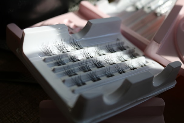 A hand holding a delicate set of natural-looking false eyelashes against a soft pastel background.