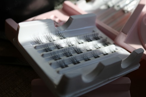 False eyelashes are neatly arranged in a plastic case with a few individual lashes missing. The case and surrounding area are lit by natural light creating some shadows.