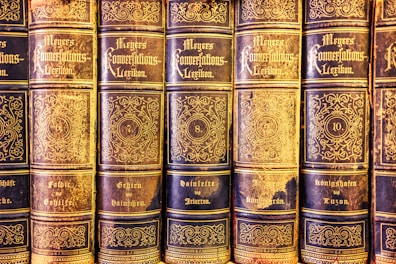 A collection of antique books lined up in a row, featuring ornate gold lettering and intricate designs on the spines. The volumes are part of 'Meyers Konversations-Lexikon,' indicated by the decorative script. Each book is numbered, with visible numbering including 6, 7, 8, and 10, and topics covered on the books' spines, such as 'Geheim' and 'Königshofen.' The books exhibit a classic and vintage aesthetic with evident signs of aging and wear.