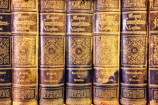 A collection of antique books lined up in a row, featuring ornate gold lettering and intricate designs on the spines. The volumes are part of 'Meyers Konversations-Lexikon,' indicated by the decorative script. Each book is numbered, with visible numbering including 6, 7, 8, and 10, and topics covered on the books' spines, such as 'Geheim' and 'Königshofen.' The books exhibit a classic and vintage aesthetic with evident signs of aging and wear.