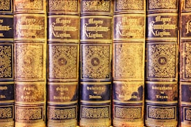 A collection of antique books lined up in a row, featuring ornate gold lettering and intricate designs on the spines. The volumes are part of 'Meyers Konversations-Lexikon,' indicated by the decorative script. Each book is numbered, with visible numbering including 6, 7, 8, and 10, and topics covered on the books' spines, such as 'Geheim' and 'Königshofen.' The books exhibit a classic and vintage aesthetic with evident signs of aging and wear.