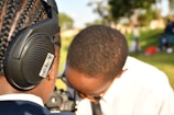 A person with braided hair is wearing large headphones with a visible barcode sticker. Another person is using a camera or some equipment, with their face partially hidden. The background is an outdoor setting, possibly a park, with green grass and trees.
