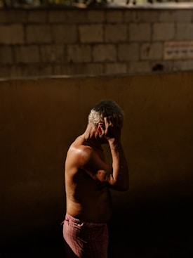 A mature man stands in profile, shirtless, against a textured wall made of concrete blocks. He is holding his head with one hand, casting a distinct shadow that highlights the texture of his skin and the muscles in his arm. The lighting emphasizes the contours of his body, creating a dramatic contrast between light and shadow.