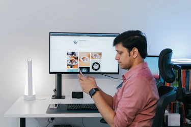 Indian marketer checking his phone at a desk, with a social media page on the monitor