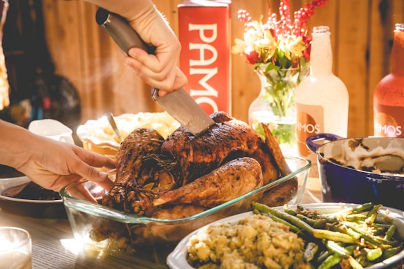A Thanksgiving or festive meal being prepared with a hand carving a roasted turkey. Surrounding the turkey, there are dishes of mashed potatoes, green beans, and stuffing. There are also bottles, a vase of flowers, and the steam indicates the food is warm.