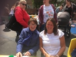 Several people are gathered outdoors, enjoying a social event. Two women are seated in front, smiling at the camera, while others stand behind them. They are dressed casually, and there is a baby stroller and a picnic table with colorful items on it in the background.
