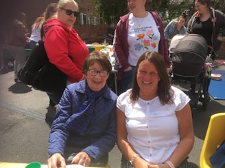 Several people are gathered outdoors, enjoying a social event. Two women are seated in front, smiling at the camera, while others stand behind them. They are dressed casually, and there is a baby stroller and a picnic table with colorful items on it in the background.