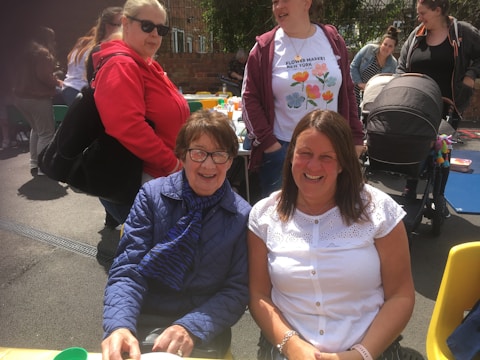 Several people are gathered outdoors, enjoying a social event. Two women are seated in front, smiling at the camera, while others stand behind them. They are dressed casually, and there is a baby stroller and a picnic table with colorful items on it in the background.