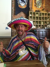 A smiling person is wearing a colorful sombrero and a poncho with horizontal stripes. The individual is giving thumbs up and has a name tag with an animal illustration. The background features banners with crests and various decorative items on shelves against a brick wall.