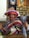 A smiling person is wearing a colorful sombrero and a poncho with horizontal stripes. The individual is giving thumbs up and has a name tag with an animal illustration. The background features banners with crests and various decorative items on shelves against a brick wall.