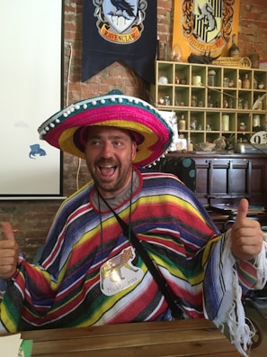 A smiling person is wearing a colorful sombrero and a poncho with horizontal stripes. The individual is giving thumbs up and has a name tag with an animal illustration. The background features banners with crests and various decorative items on shelves against a brick wall.