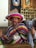 A smiling person is wearing a colorful sombrero and a poncho with horizontal stripes. The individual is giving thumbs up and has a name tag with an animal illustration. The background features banners with crests and various decorative items on shelves against a brick wall.