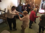 A group of elderly people are engaging in a dance activity in a room. Some chairs are stacked on the side, and a piano is visible in the background. The atmosphere appears lively and social.