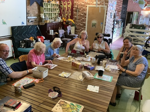 A group of people, mostly older adults, sits around a large wooden table in a cozy room with exposed brick walls. They are engaged in a craft or gaming activity with various items spread across the table including papers, boxes, mugs, and crafts supplies. The room is decorated with hanging curtains, shelving with assorted items, and a variety of other decorations.