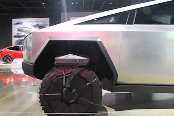 A futuristic vehicle with a geometric design and large, rugged wheels. The body is metallic with angular shapes, conveying strength and modernity. In the background, there is a red car and automotive designs displayed on posters.