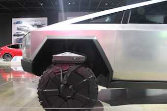 A futuristic vehicle with a geometric design and large, rugged wheels. The body is metallic with angular shapes, conveying strength and modernity. In the background, there is a red car and automotive designs displayed on posters.