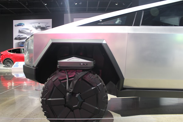 A futuristic vehicle with a geometric design and large, rugged wheels. The body is metallic with angular shapes, conveying strength and modernity. In the background, there is a red car and automotive designs displayed on posters.