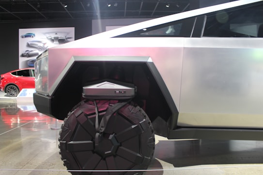 A futuristic vehicle with a geometric design and large, rugged wheels. The body is metallic with angular shapes, conveying strength and modernity. In the background, there is a red car and automotive designs displayed on posters.