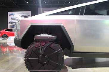 A futuristic vehicle with a geometric design and large, rugged wheels. The body is metallic with angular shapes, conveying strength and modernity. In the background, there is a red car and automotive designs displayed on posters.