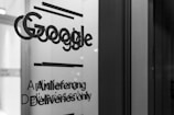 A grayscale photo showing the word 'Google' along with text in English and German on a glass surface, possibly a door or window. The surrounding area appears out of focus, emphasizing the text.