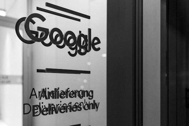 A grayscale photo showing the word 'Google' along with text in English and German on a glass surface, possibly a door or window. The surrounding area appears out of focus, emphasizing the text.