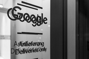 A grayscale photo showing the word 'Google' along with text in English and German on a glass surface, possibly a door or window. The surrounding area appears out of focus, emphasizing the text.