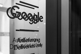 A grayscale photo showing the word 'Google' along with text in English and German on a glass surface, possibly a door or window. The surrounding area appears out of focus, emphasizing the text.