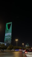 A nighttime cityscape with a tall, modern skyscraper illuminated in green lights and displaying Arabic text. The foreground includes a road with vehicles and streetlights casting a warm glow, while trees line the streets.