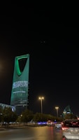 A nighttime cityscape with a tall, modern skyscraper illuminated in green lights and displaying Arabic text. The foreground includes a road with vehicles and streetlights casting a warm glow, while trees line the streets.