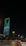 A nighttime cityscape with a tall, modern skyscraper illuminated in green lights and displaying Arabic text. The foreground includes a road with vehicles and streetlights casting a warm glow, while trees line the streets.