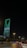 A nighttime cityscape with a tall, modern skyscraper illuminated in green lights and displaying Arabic text. The foreground includes a road with vehicles and streetlights casting a warm glow, while trees line the streets.