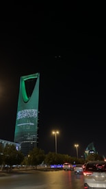 A nighttime cityscape with a tall, modern skyscraper illuminated in green lights and displaying Arabic text. The foreground includes a road with vehicles and streetlights casting a warm glow, while trees line the streets.