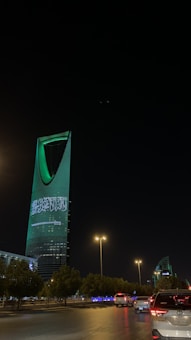 A nighttime cityscape with a tall, modern skyscraper illuminated in green lights and displaying Arabic text. The foreground includes a road with vehicles and streetlights casting a warm glow, while trees line the streets.