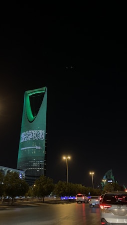A nighttime cityscape with a tall, modern skyscraper illuminated in green lights and displaying Arabic text. The foreground includes a road with vehicles and streetlights casting a warm glow, while trees line the streets.
