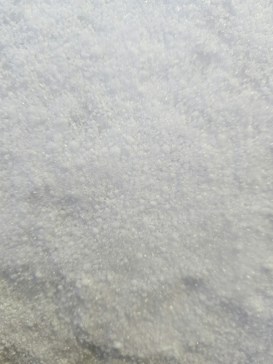 A close-up view of a surface covered in fine, white granular material that appears similar to snow or sugar, with a slightly glistening texture.