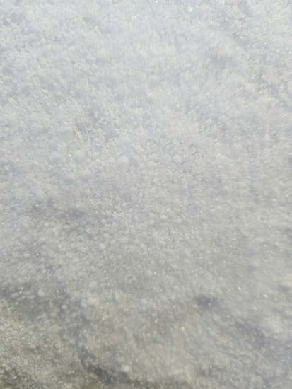 A close-up view of a surface covered in fine, white granular material that appears similar to snow or sugar, with a slightly glistening texture.