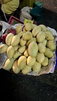 A pile of ripe, yellow mangoes placed on a paper-lined surface, with a handwritten price card displaying the number 50. In the background, there are plastic baskets and containers, highlighting a rustic market setting.