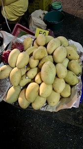 A pile of ripe, yellow mangoes placed on a paper-lined surface, with a handwritten price card displaying the number 50. In the background, there are plastic baskets and containers, highlighting a rustic market setting.